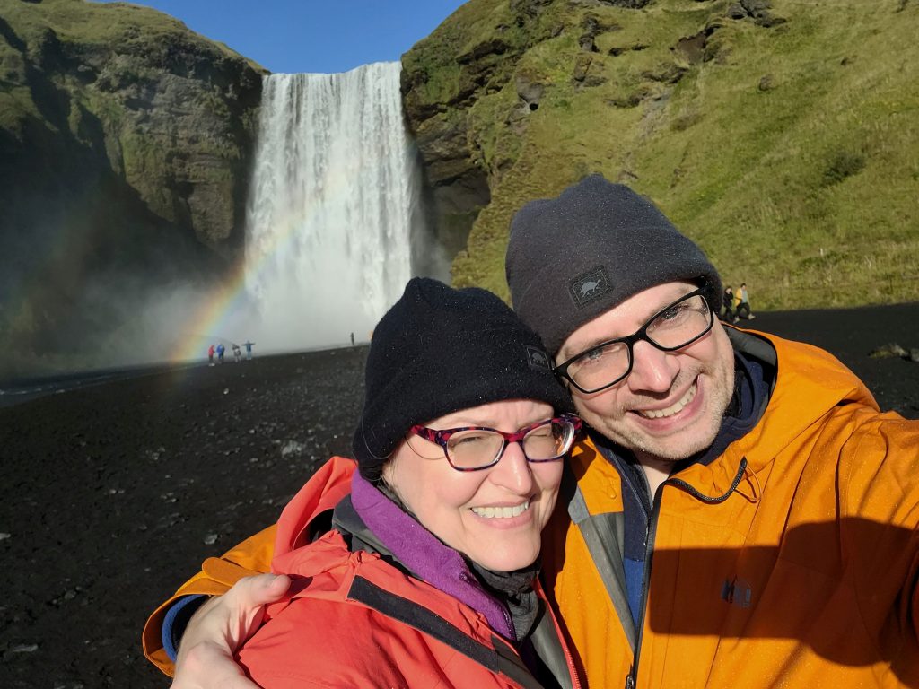 Susan and Graham with a rainbow, waterfall, and mountain behind them.