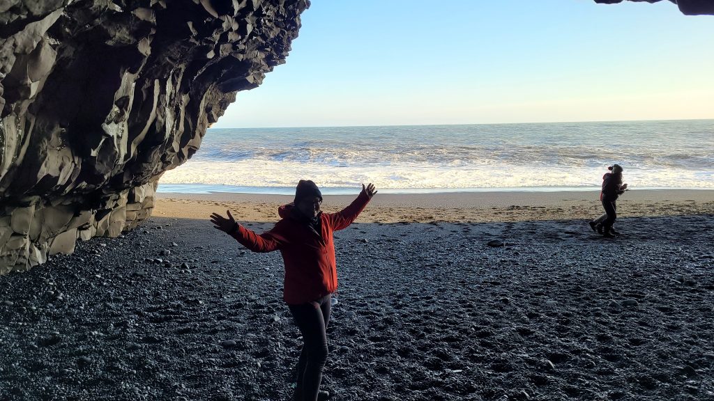 Susan standing inside a basalt sea cave with black sands and the ocean in the background