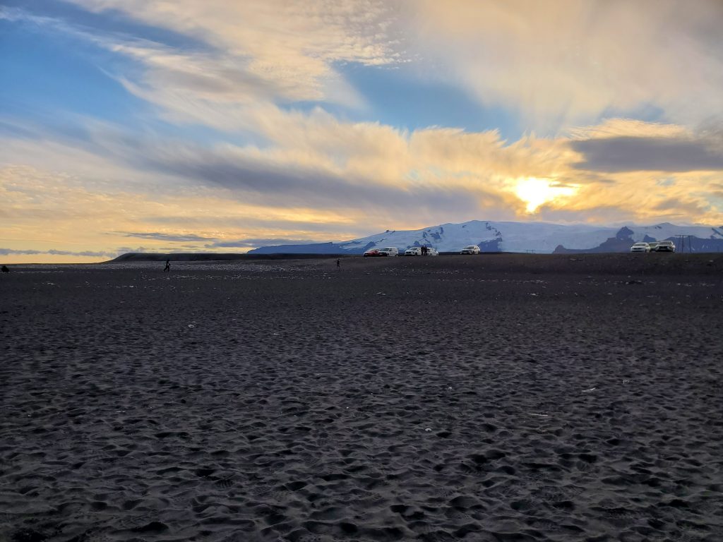 parking lot at top of black sand beach with glacier in the backround and low sun with clouds